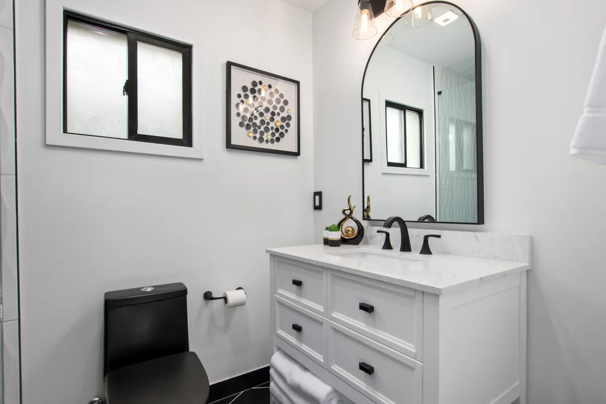 A clean and contemporary bathroom featuring a vanity with a marble countertop, round mirror, black fixtures, and elegant wall art. The setting feels serene and tailored for self-care routines.