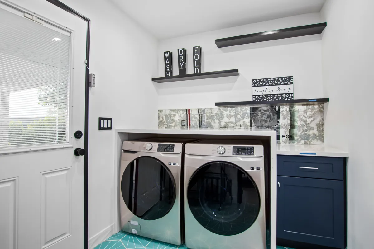 A utility area with a washer and dryer set against blue cabinetry and open shelving with decorative elements. The space looks organized and clean, ideal for handling family laundry and daily routines.