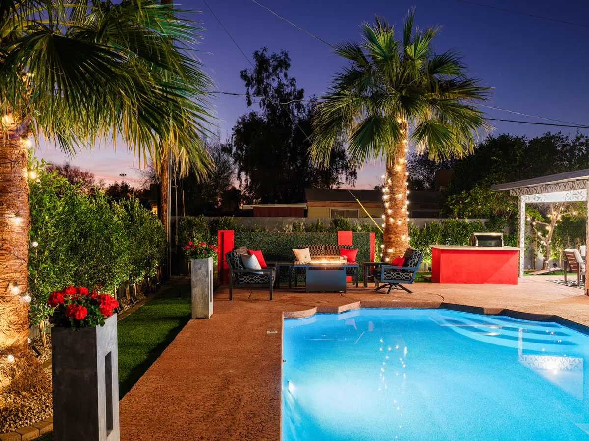 A beautifully arranged backyard with a sparkling swimming pool, palm trees, fire pit, and a barbecue area. Comfortable seating invites guests to gather, dine, and share laughter well into the evening.