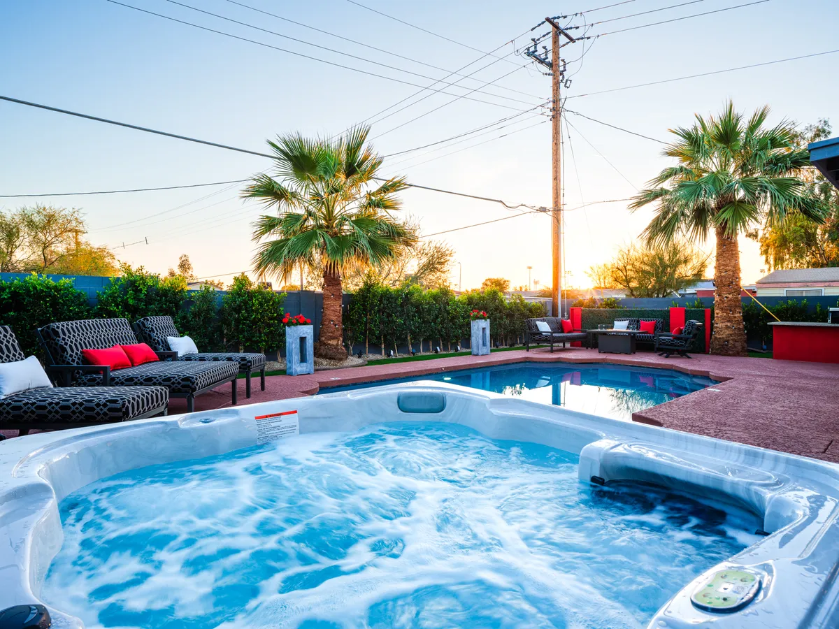 This image showcases a sparkling swimming pool with an adjacent hot tub, surrounded by palm trees and inviting outdoor seating. The view is set at sunset, creating a warm, relaxing atmosphere. The setup reflects both the luxury and fun of a resort-style backyard.
