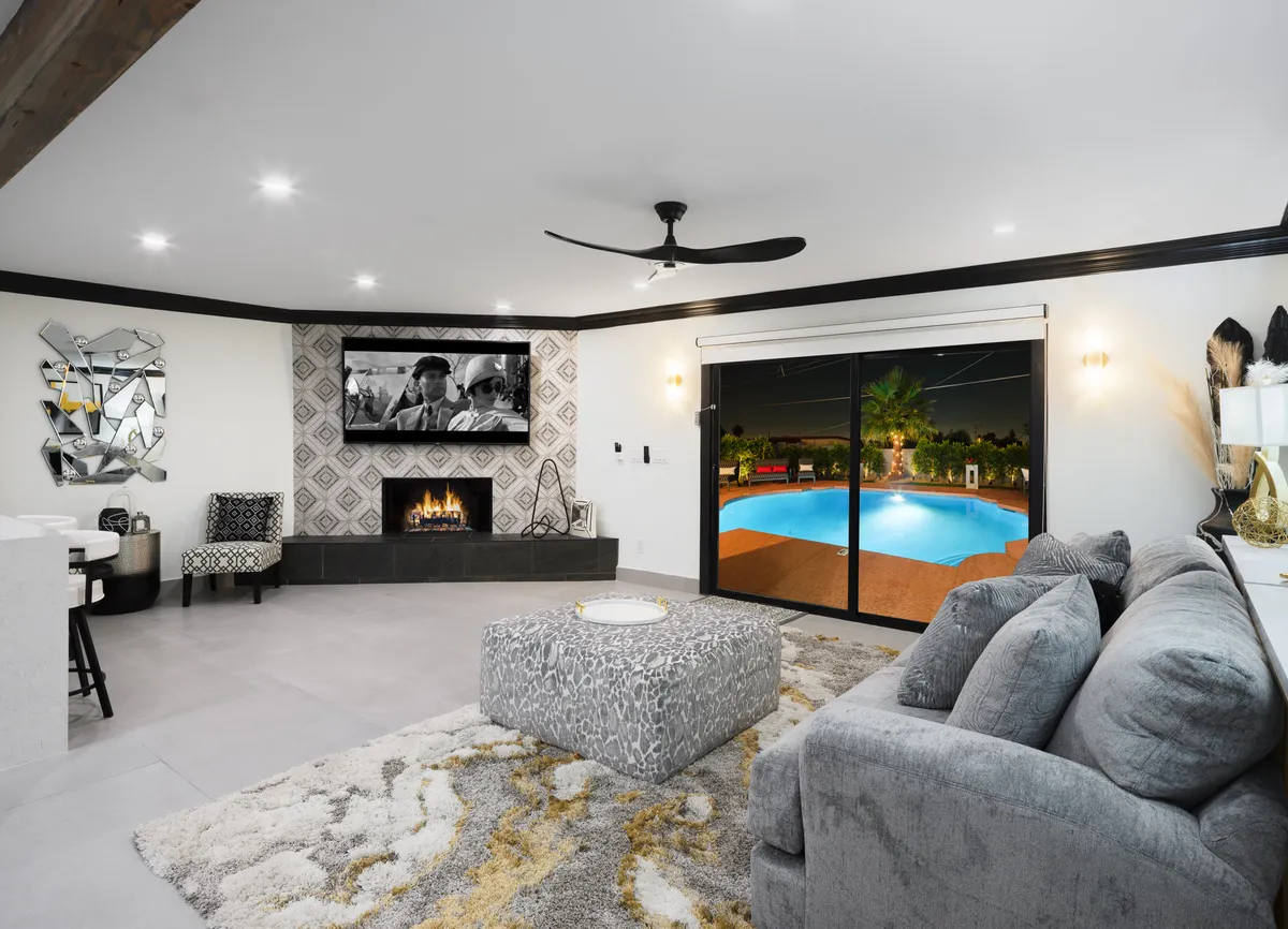 This image features a stylish living room anchored by a glowing fireplace and a television above it. There’s a comfortable seating area arranged around the fire, decorative patterns, and a sliding glass door offering a glimpse of the pool outside. The ambiance is cozy yet modern, perfect for socializing or relaxing.