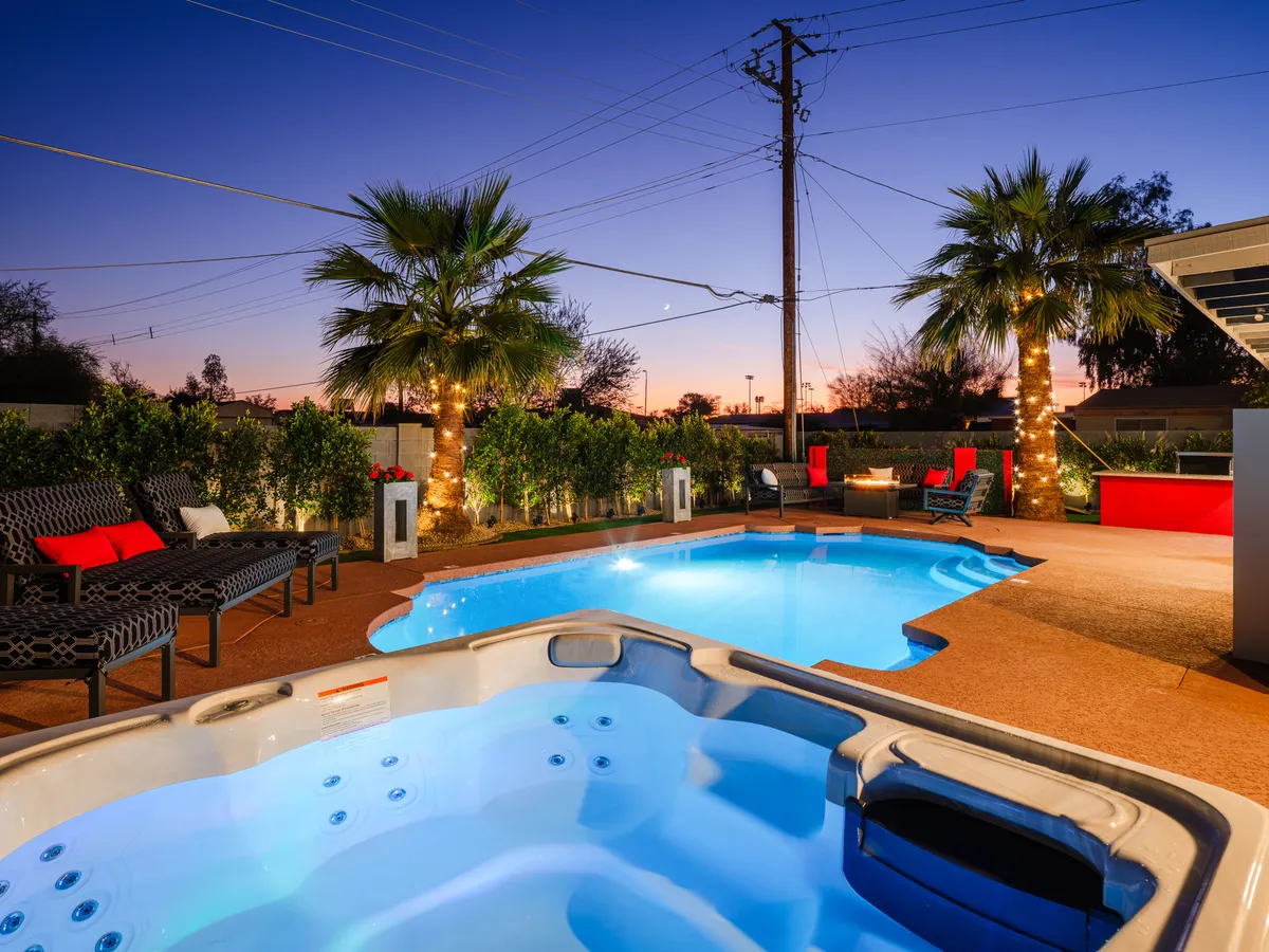 A festive backyard scene with a sparkling swimming pool, bubbling hot tub, palm trees, and a spacious outdoor seating area. The space is clearly designed for relaxation and group gatherings, with ample room for socializing under the stars.