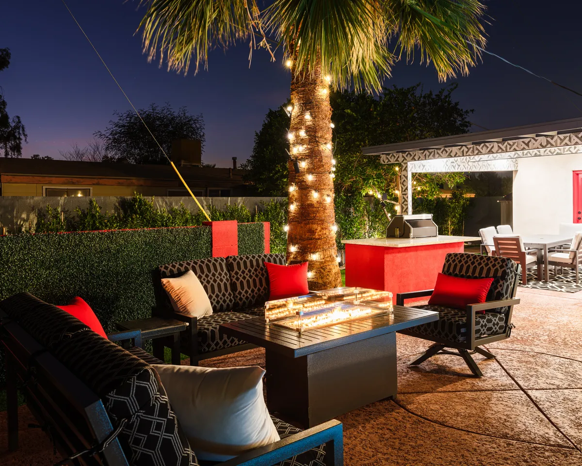 A palm tree adorned with string lights towers over a glowing fire pit, with a spacious outdoor seating area and a BBQ grill visible in the background. The scene is set for evening relaxation or lively social gatherings.