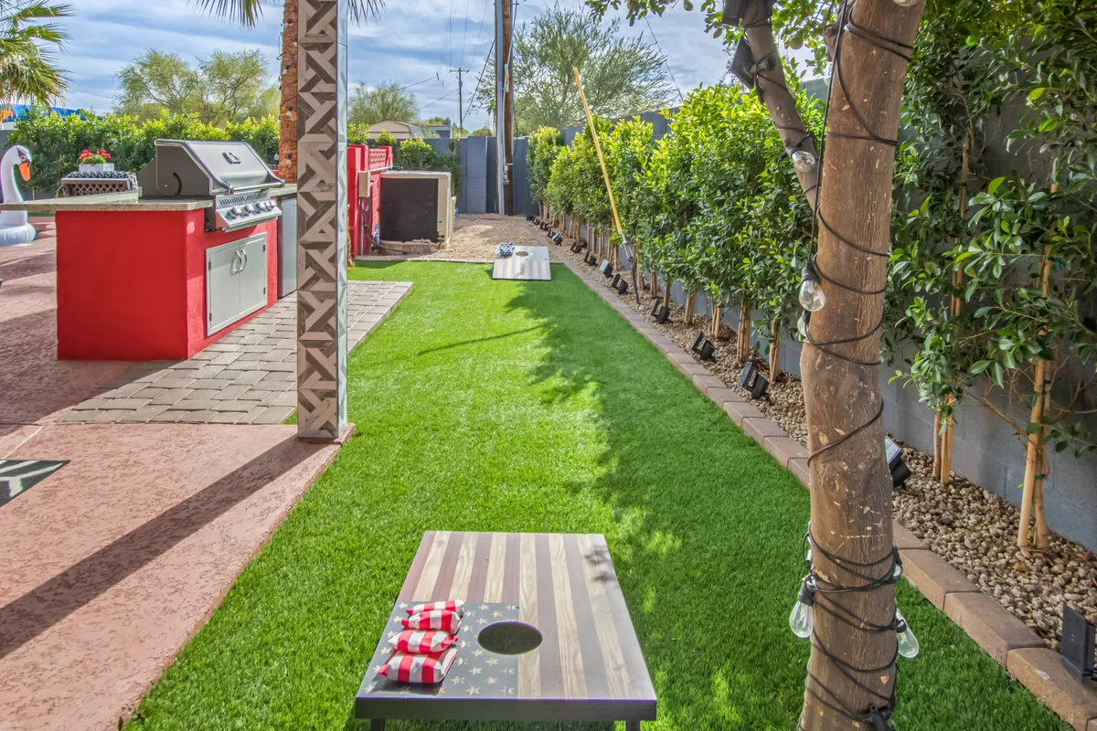 A cornhole board rests on a lush green lawn next to a barbecue grill, with a lighted tree casting a festive glow. The open space invites outdoor games, cookouts, and playful competitions.