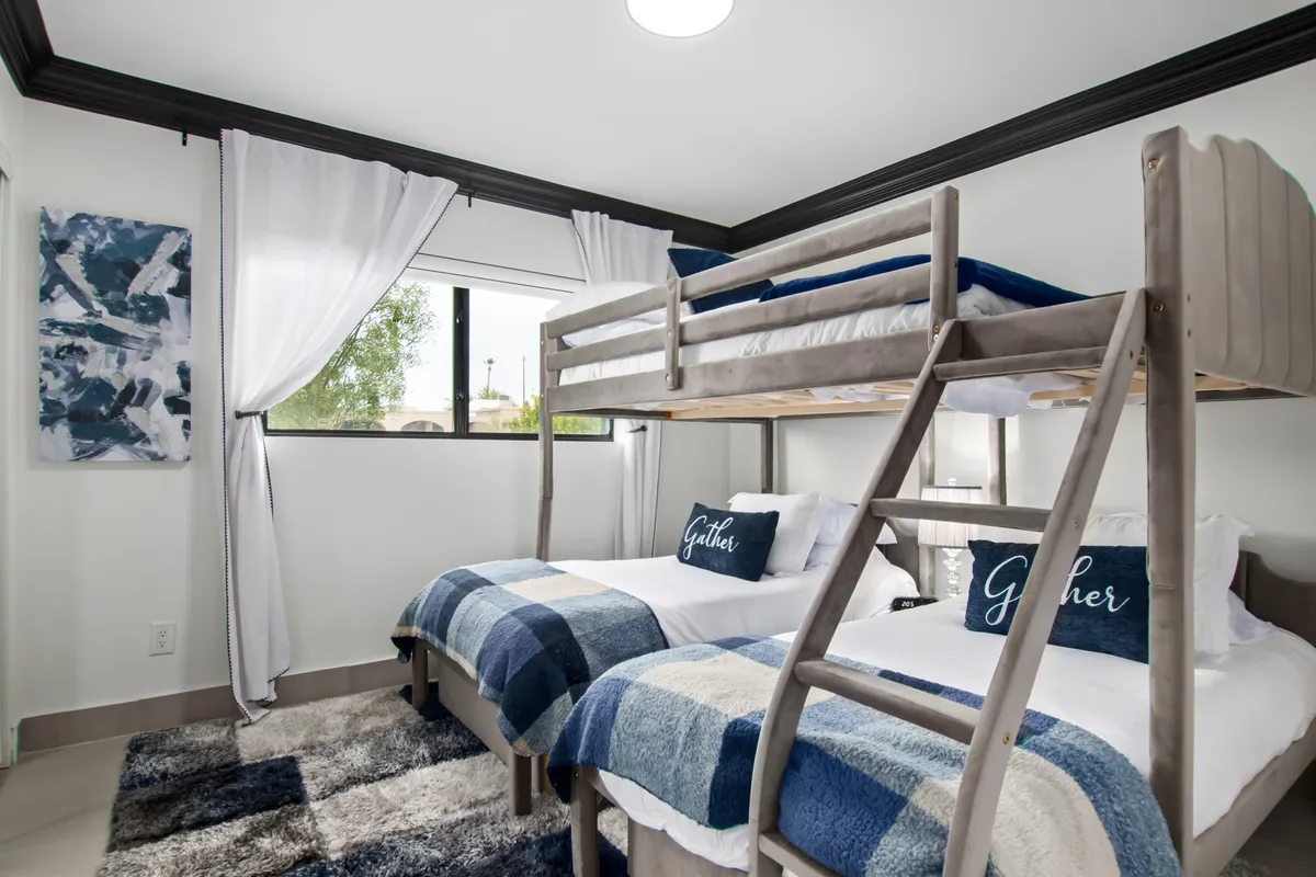 A playful, inviting bunk bedroom with a full-over-twin bunk bed, plaid blankets, decorative throw pillows, and a window with sheer curtains. The space feels cozy and perfect for kids or friends.