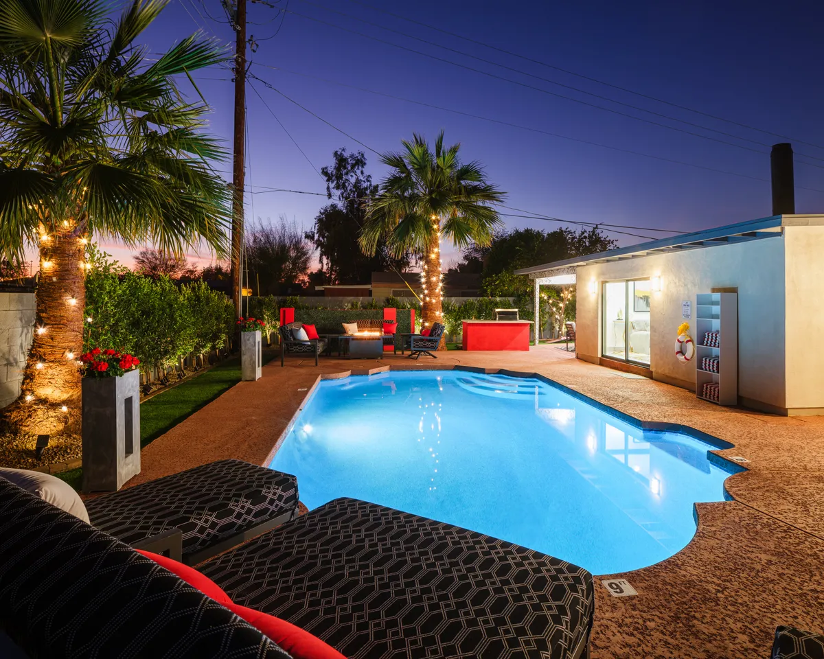 This exterior image captures a luxurious backyard with a sparkling swimming pool, surrounded by palm trees, plush lounge chairs, a fire pit, and a modern outdoor seating area. The inviting setup highlights a festive yet relaxed atmosphere for both daytime and evening enjoyment.